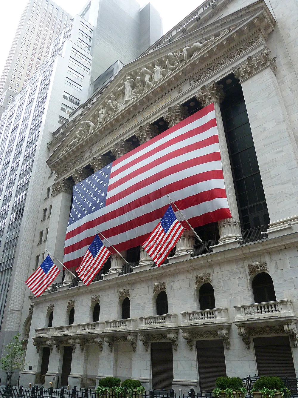 The New York Stock Exchange facade on Wall Street with a large U.S. flag across the columns. Photo: Wikimedia Commons.